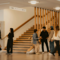 A group of people talking and walking near a wooden staircase indoors, with handrails and overhead lights visible.