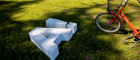 A white 3D letter 'A' sculpture on grass beside a red bicycle with a black basket in a sunny park.